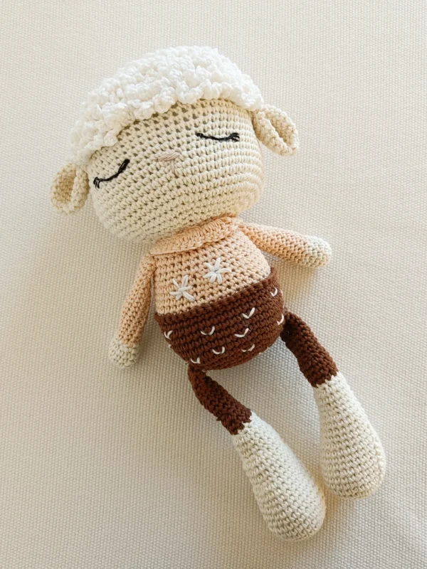 Oveja de juguete tejida a crochet (amigurumi) en tonos crema, beige y marrón, recostada sobre una superficie blanca de textura suave.