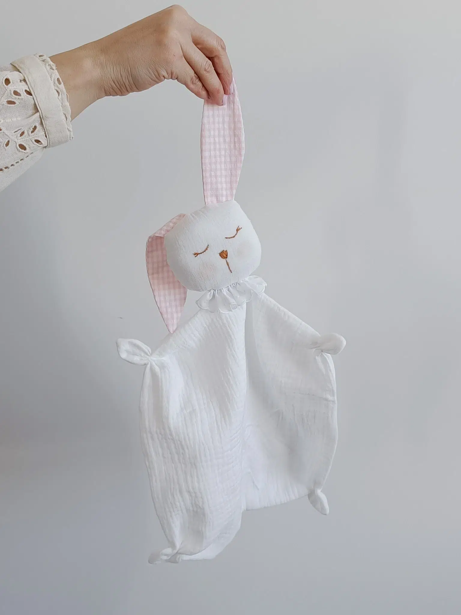 Conejito de apego para bebé de tela blanca con orejas rosadas sostenido por una mano. Conejito de apego para bebé de tela blanca con orejas rosadas sostenido por una mano.