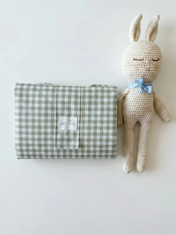 Cambiador impermeable portátil para bebé acolchado en vichy verde salvia plegado junto a conejo de ganchillo artesanal de Petit Miu