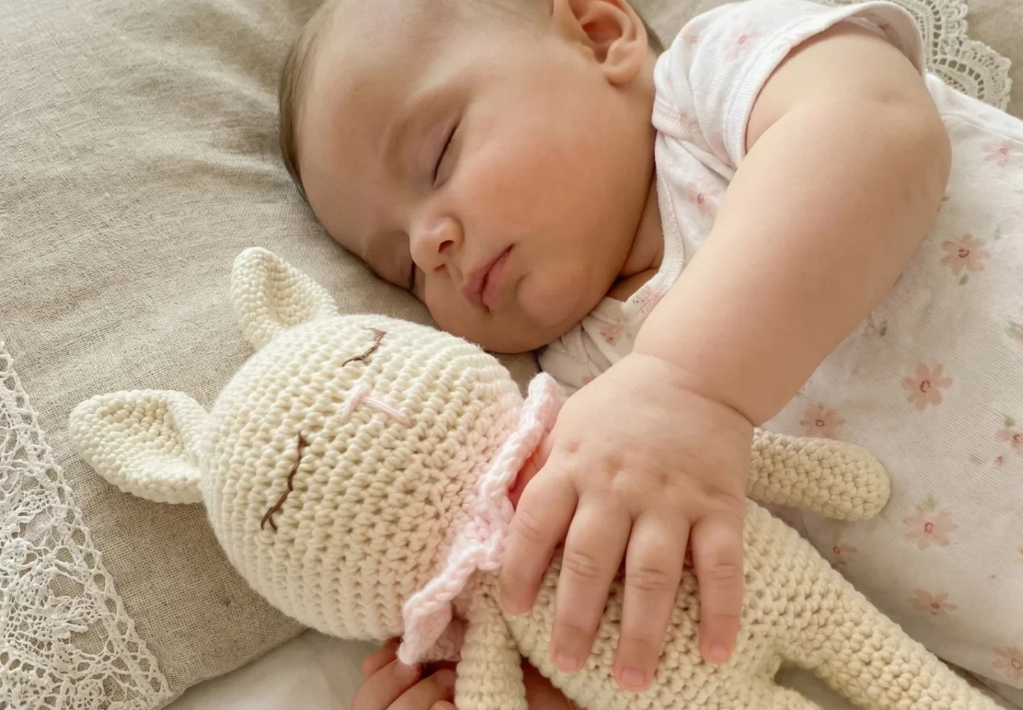 Bebé dormido abrazando un amigurumi artesanal de color crema en una cama, con mantas y encaje en el fondo. amigurumis artesanales para bebés.