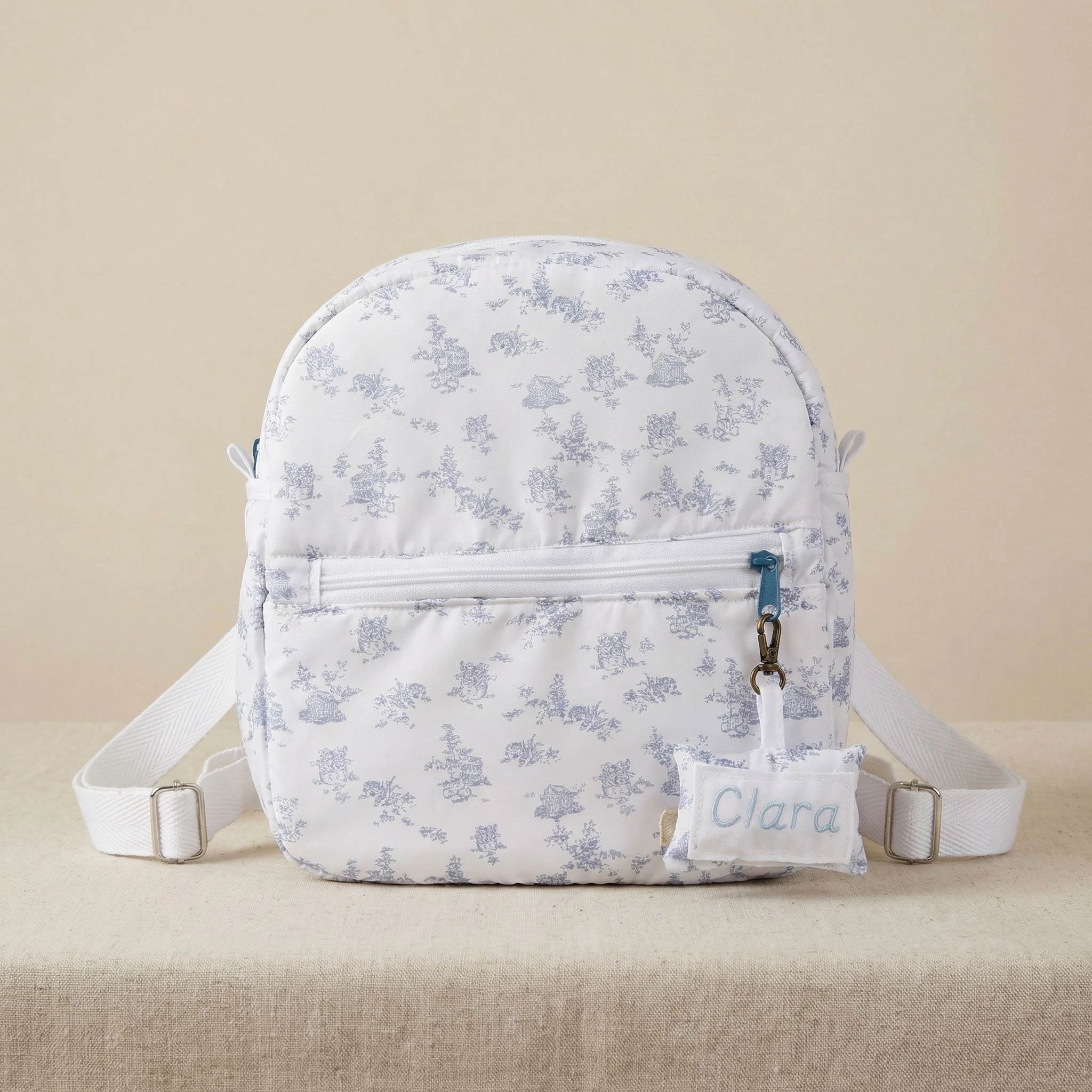 Mochila guardería personalizada blanca con bordado azul claro y diseño floral, bolsillo frontal y correa blanca adjustable.